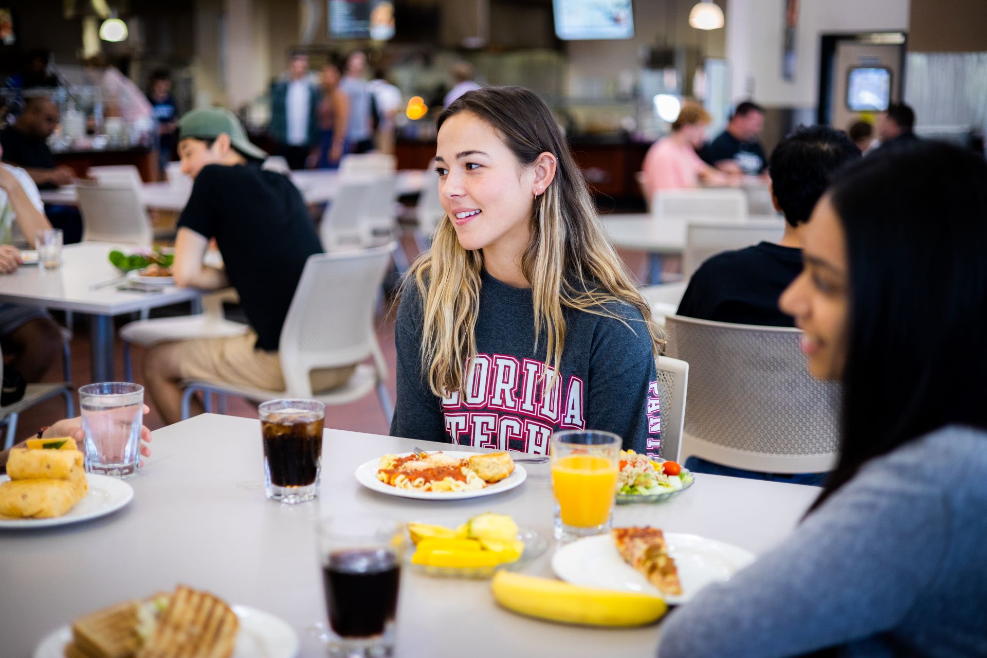 A young woman wearing a Florida Tech sweatshirt sitting at a table in a college dining hall, eating with friends
